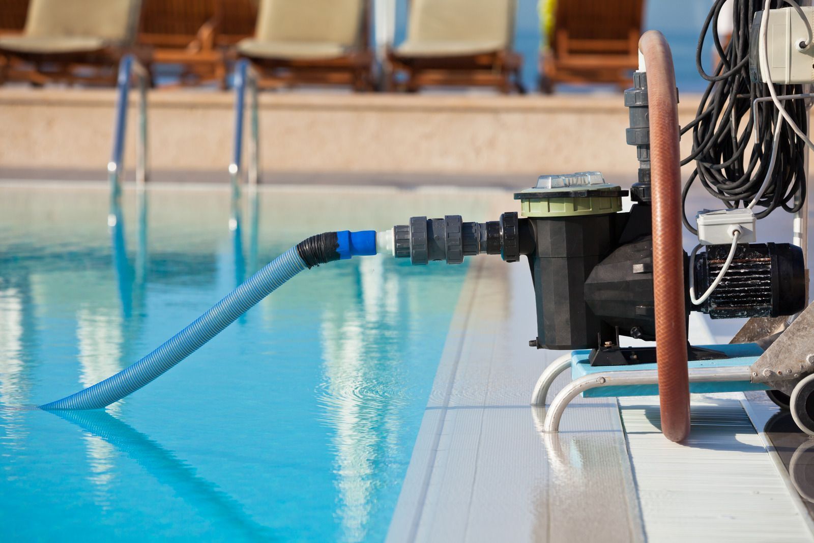 A blue pool vacuum hose connects a portable pump system to the water at the edge of an outdoor swimming pool.