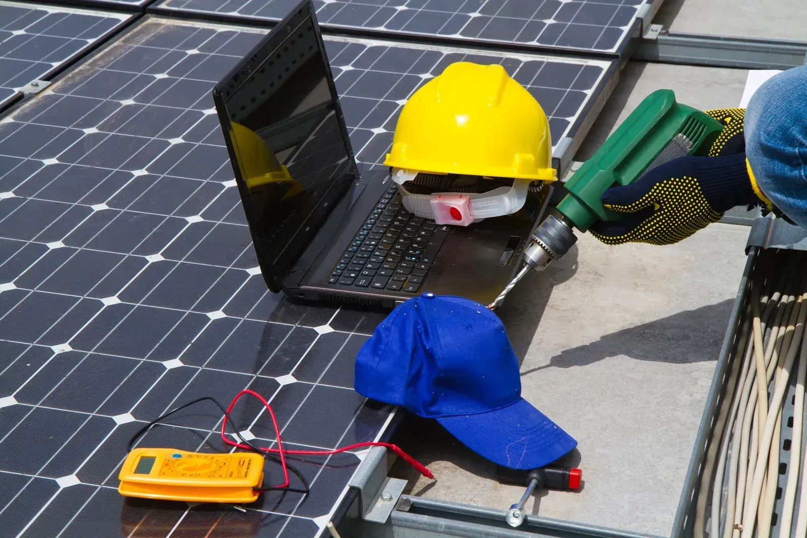 A person uses a drill near a laptop, a yellow hard hat, a blue cap, and a multimeter on a solar panel array.