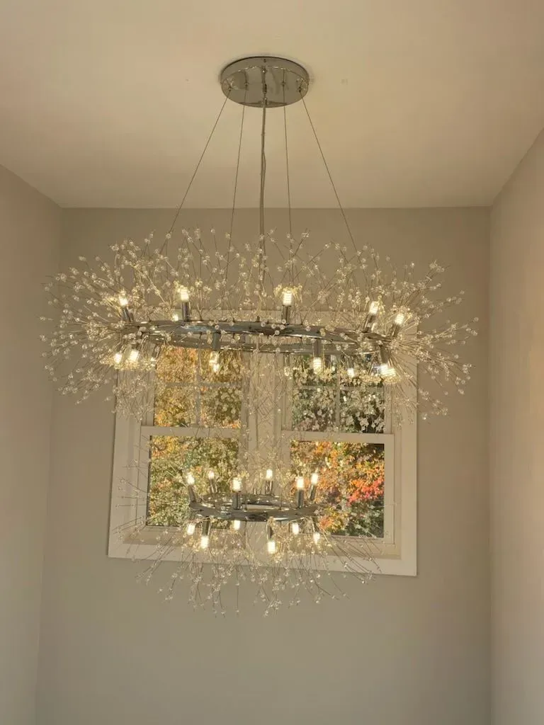 A two-tiered, circular crystal chandelier featuring metallic light spokes, suspended in front of a window.