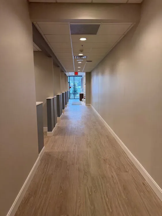 A long, brightly lit hallway with light wood-style flooring, gray partition walls on the left, and solid walls on the right.