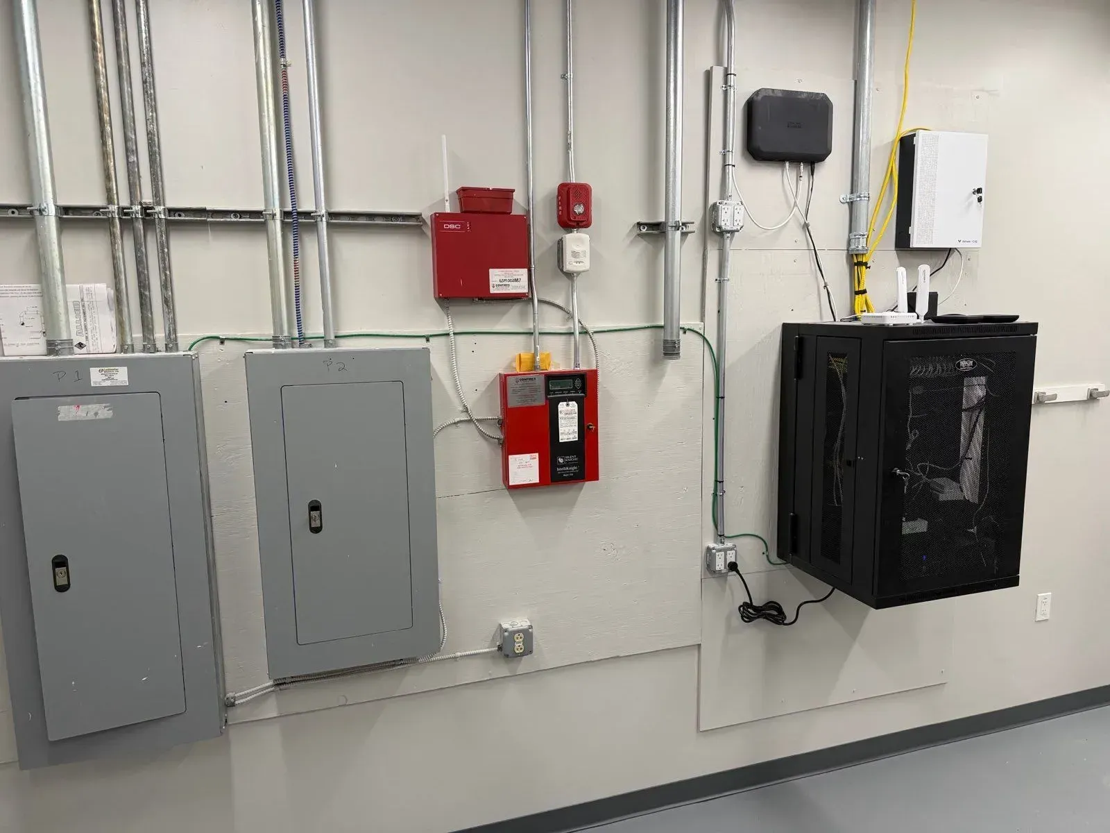 Utility wall featuring two grey electrical panels, red fire alarm control boxes, and a wall-mounted black network rack.