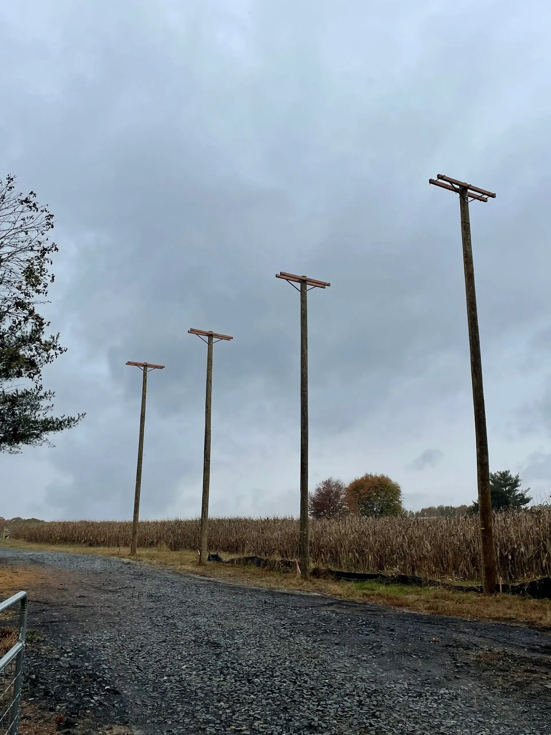 Four utility poles of increasing height stand in a line next to a gravel path and a harvested cornfield under a cloudy sky.