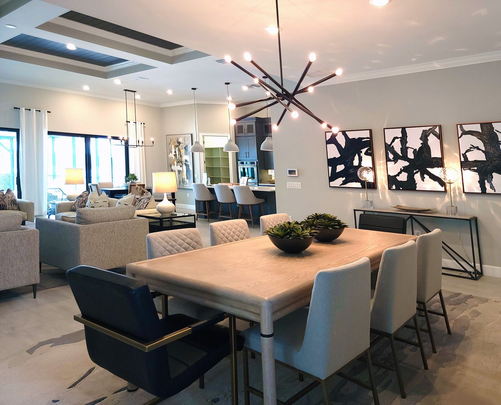 A modern dining area featuring a light wood table, upholstered chairs, a starburst chandelier, and three abstract artworks.