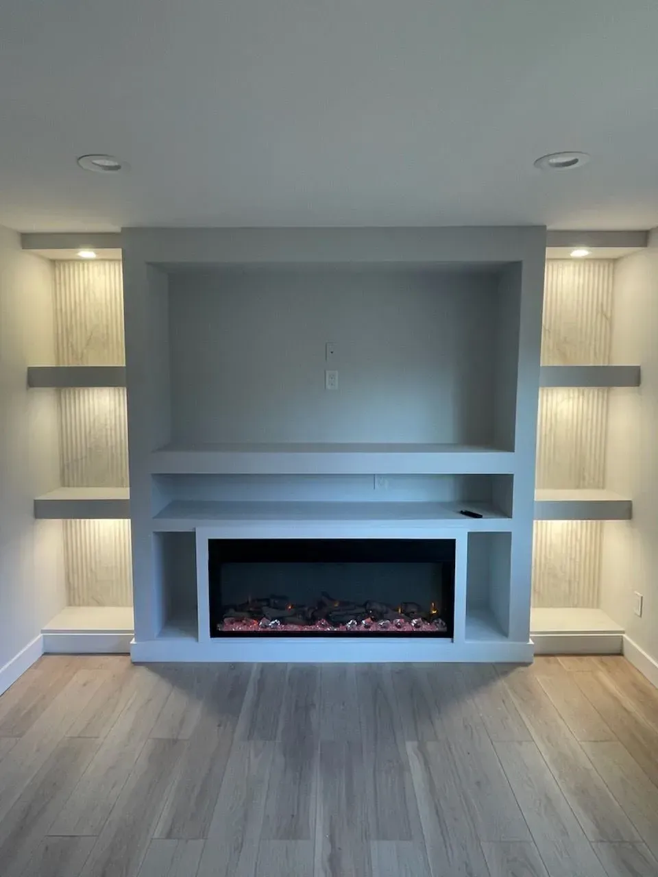 A built-in gray wall unit featuring a central electric fireplace with shelving on both sides and integrated lighting.