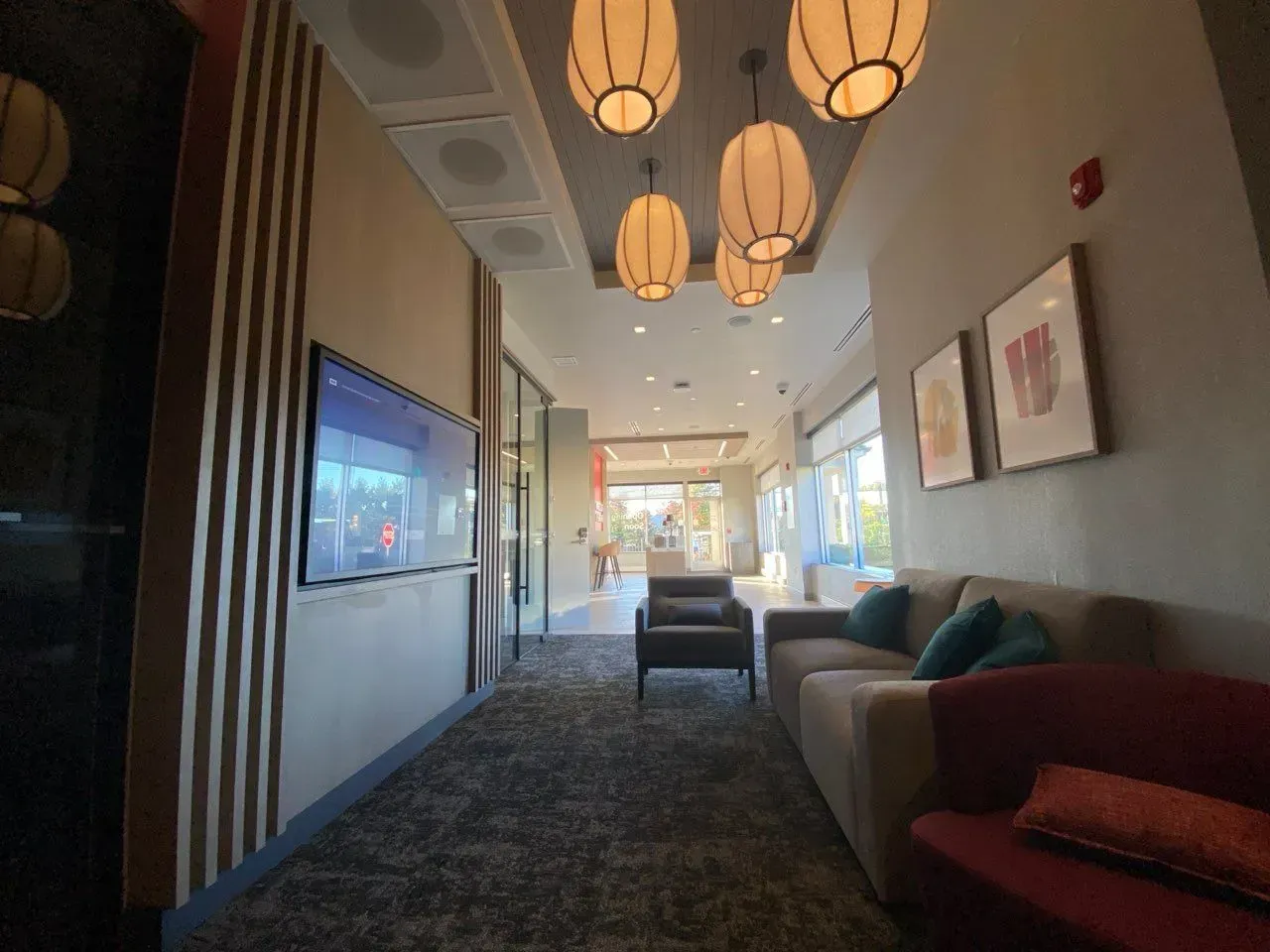 A modern lobby area with hanging lanterns, a wall-mounted TV, a gray sofa, a red chair, and framed artwork on the walls.