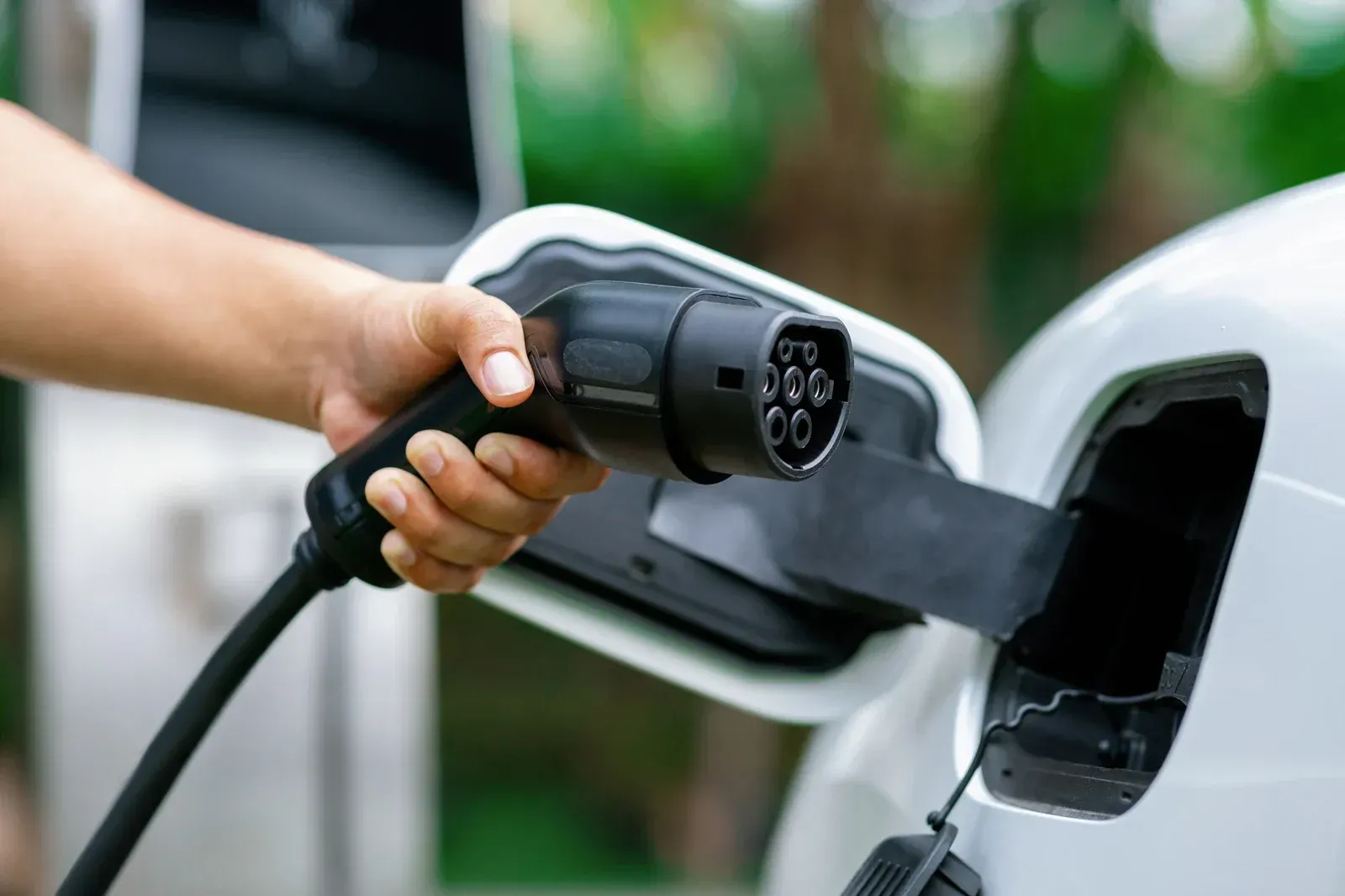 A hand holds an electric vehicle charging connector in front of the open charging port of a white car.