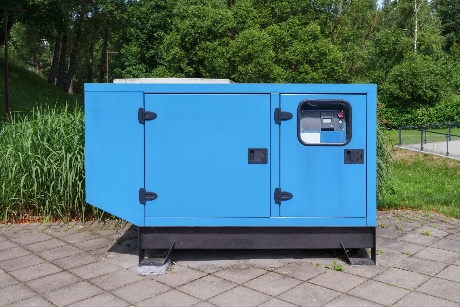 A bright blue, rectangular industrial generator sits on a paved outdoor area surrounded by green trees and grass.