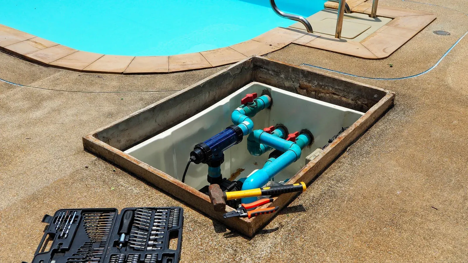 A pool plumbing box sits in the ground near a swimming pool, containing blue PVC pipes and a tool kit on the deck.