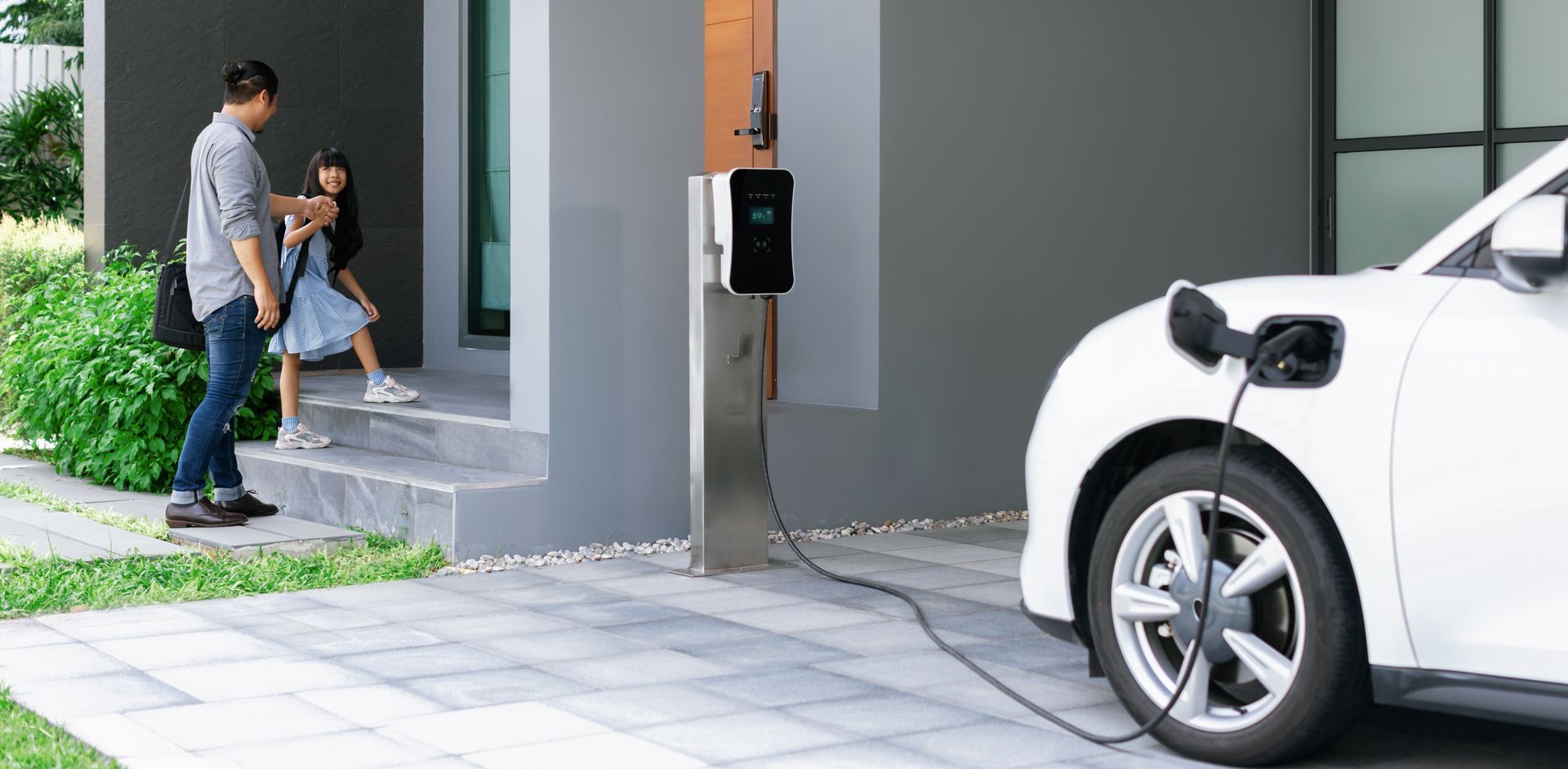 A white electric car is being charged in a driveway next to a house.