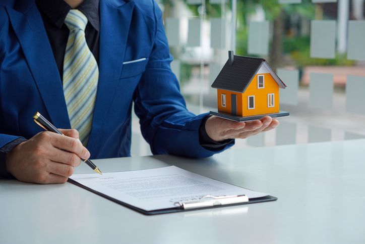 Un hombre con traje y corbata está firmando un contrato mientras sostiene una casa modelo.