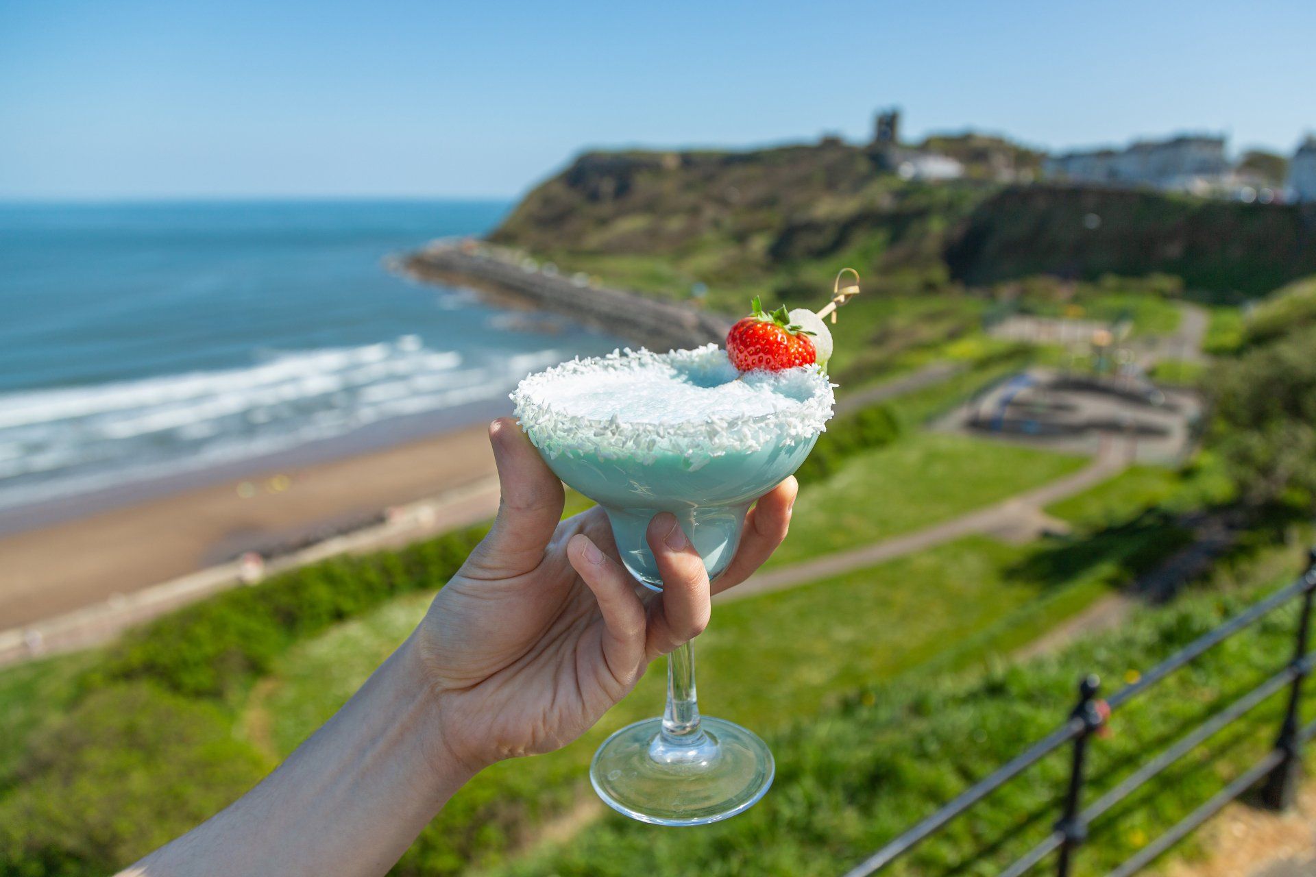 A person is holding a blue cocktail with a strawberry on top in front of the ocean.