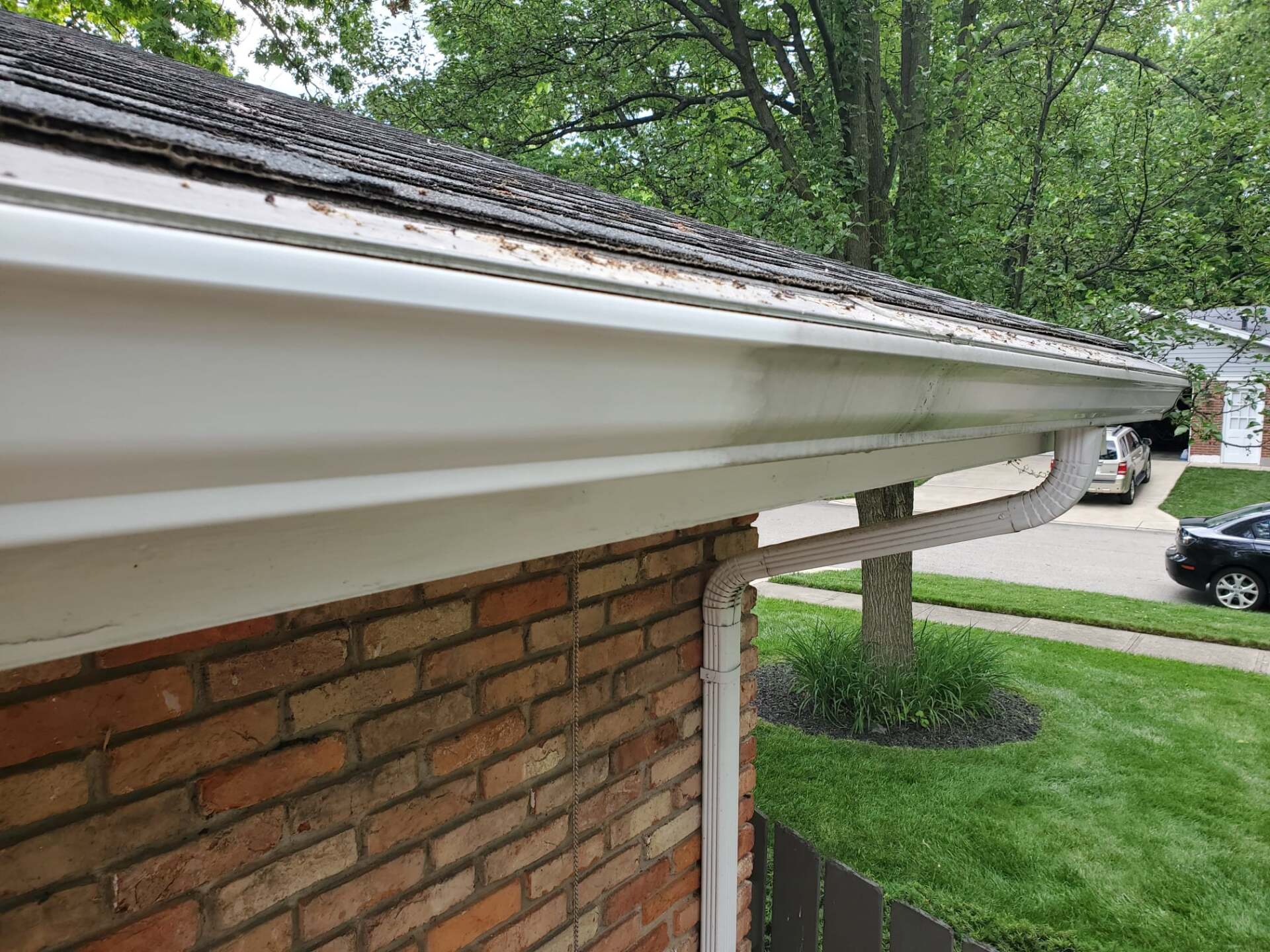 White gutters on a brick house with a downspout, green grass and a tree in the background.