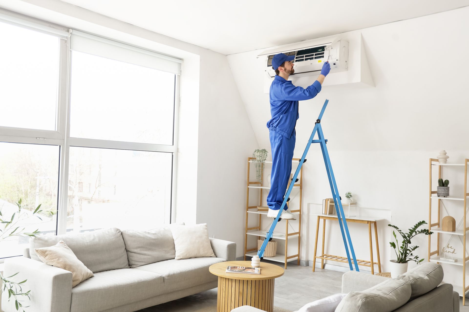 Un technicien en combinaison bleue répare un climatiseur mural dans un salon lumineux à l'aide d'un escabeau.