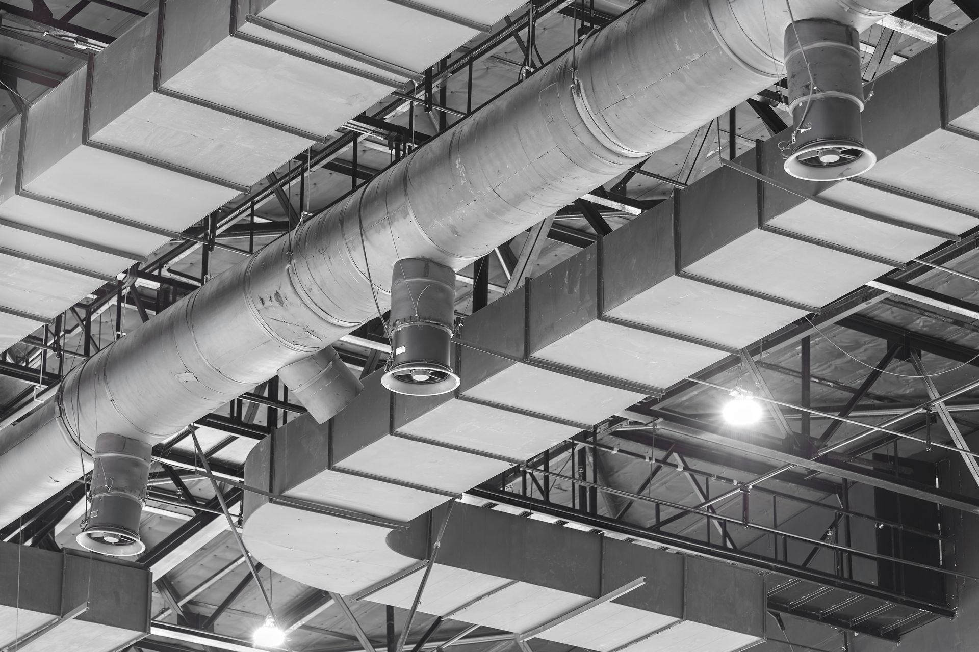 Vue du plafond montrant des conduits industriels, des fermes métalliques et des luminaires suspendus dans un grand espace intérieur