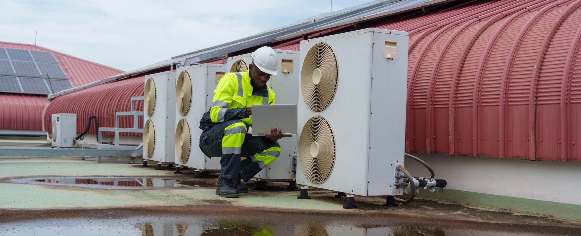 Un ouvrier en ciré jaune inspecte les unités de climatisation sur le toit d'un bâtiment rouge incurvé.
