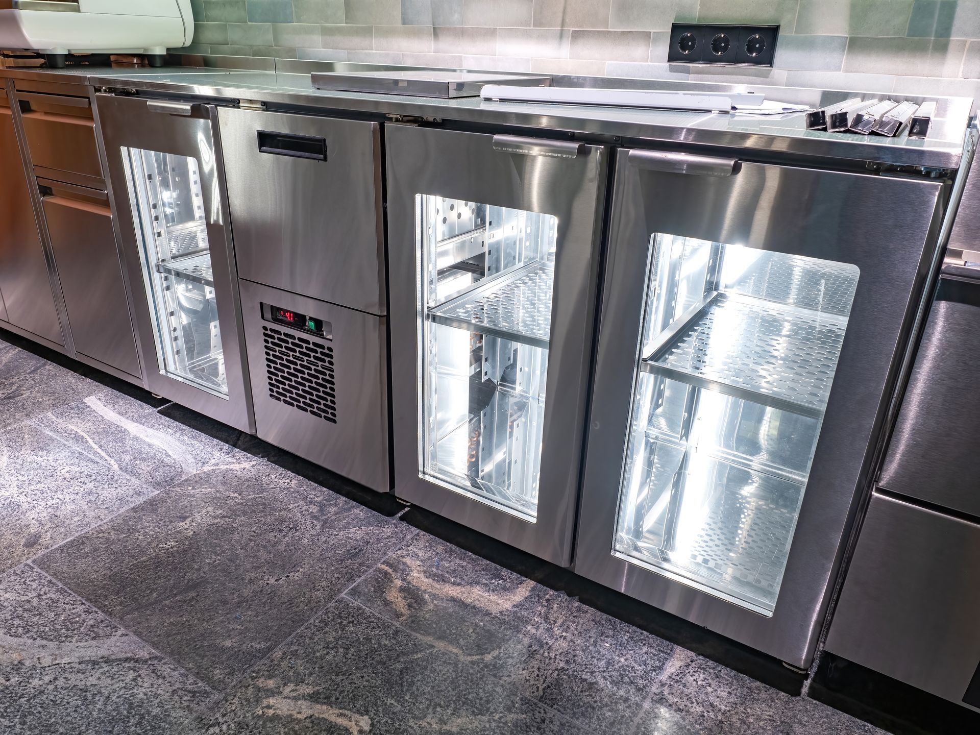 Armoires réfrigérées sous plan de travail en acier inoxydable avec portes vitrées et étagères dans une cuisine.