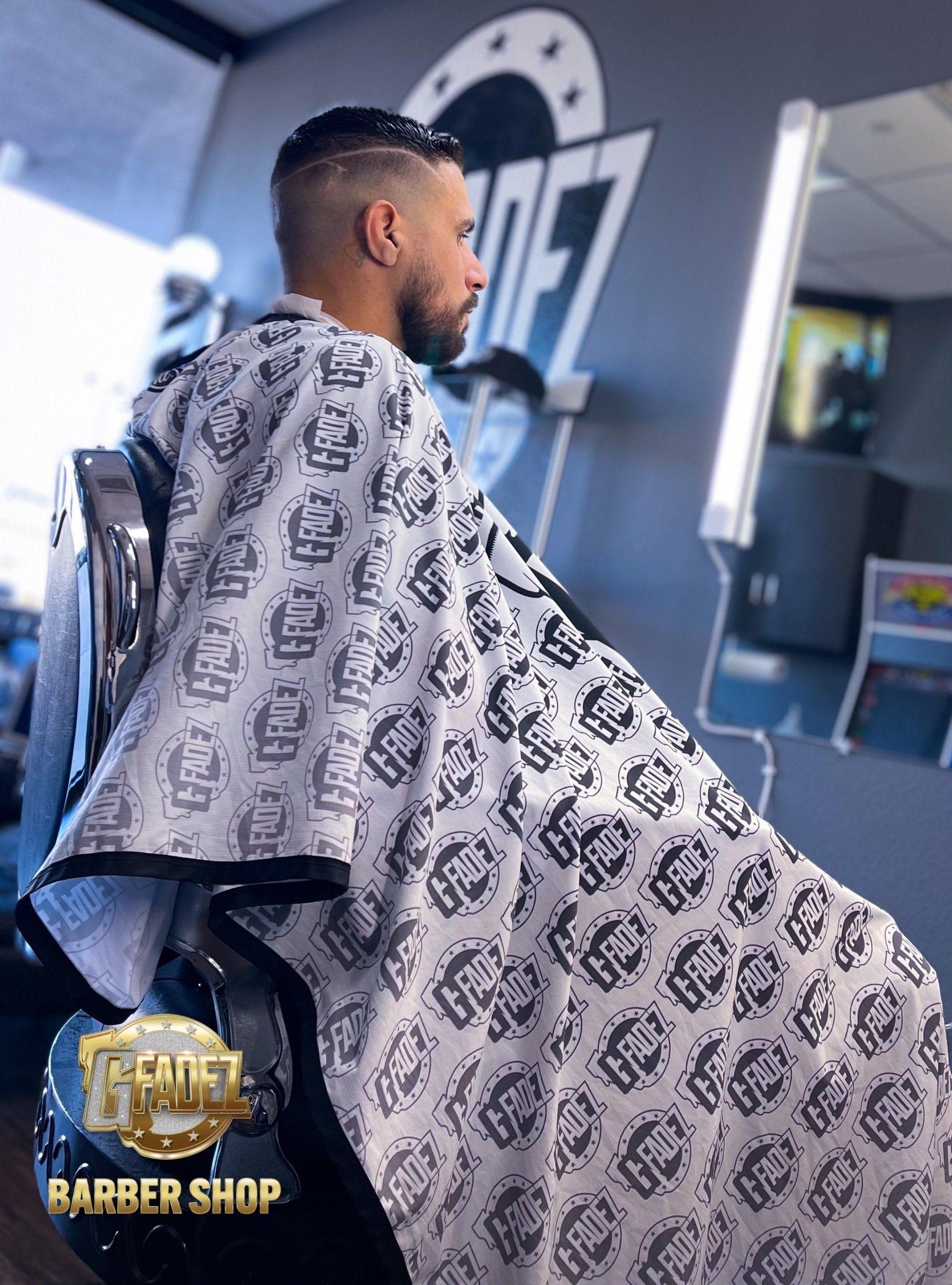 Barber shop interior with a client under a patterned cape beside a lit mirror and logo wall