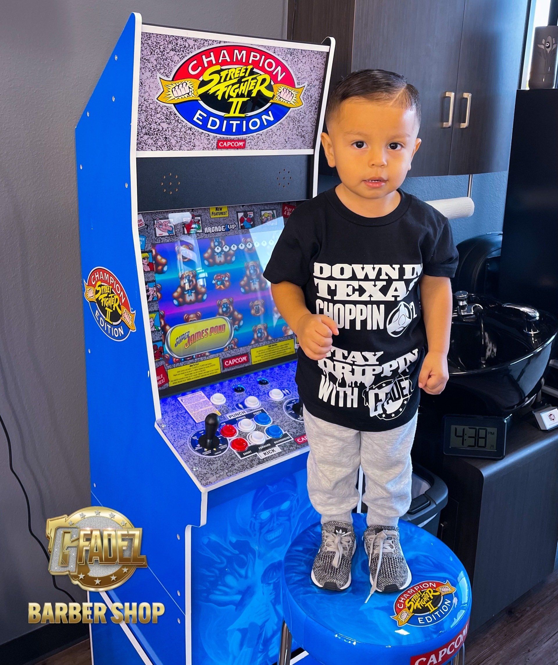 Child standing on a blue arcade machine in a barbershop, wearing a black shirt and gray pants.
