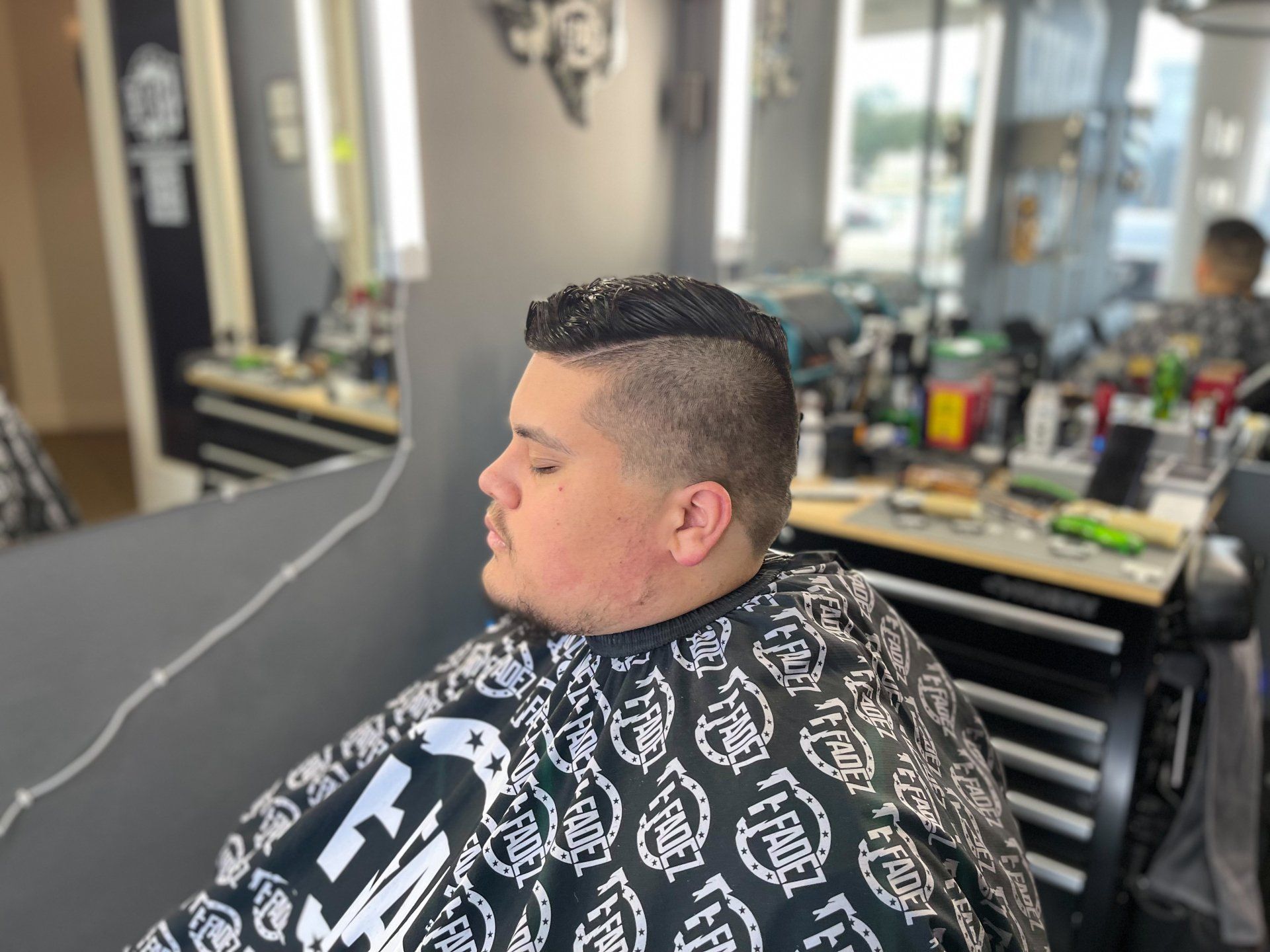 Person seated in a barber chair wearing a black cape, with a short fade haircut, in a barbershop