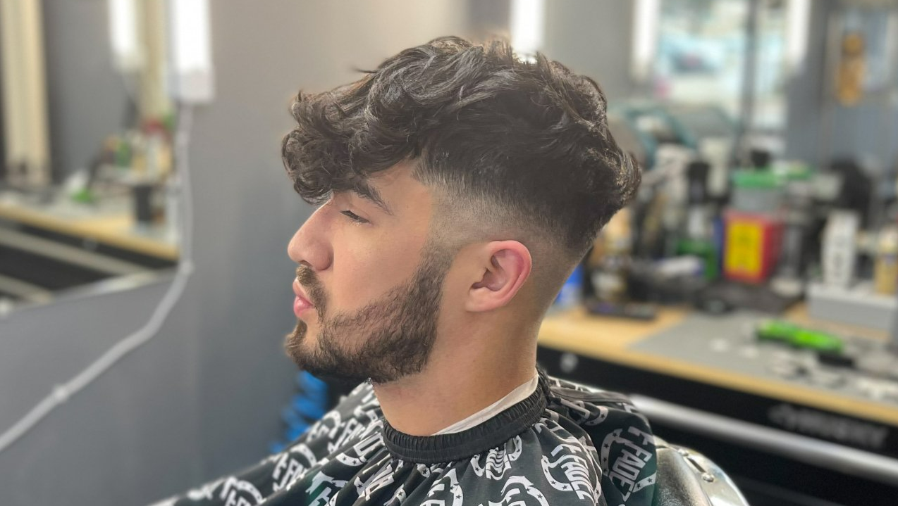 Side-profile haircut at a barbershop, showing a man with curly top fade and trimmed beard.