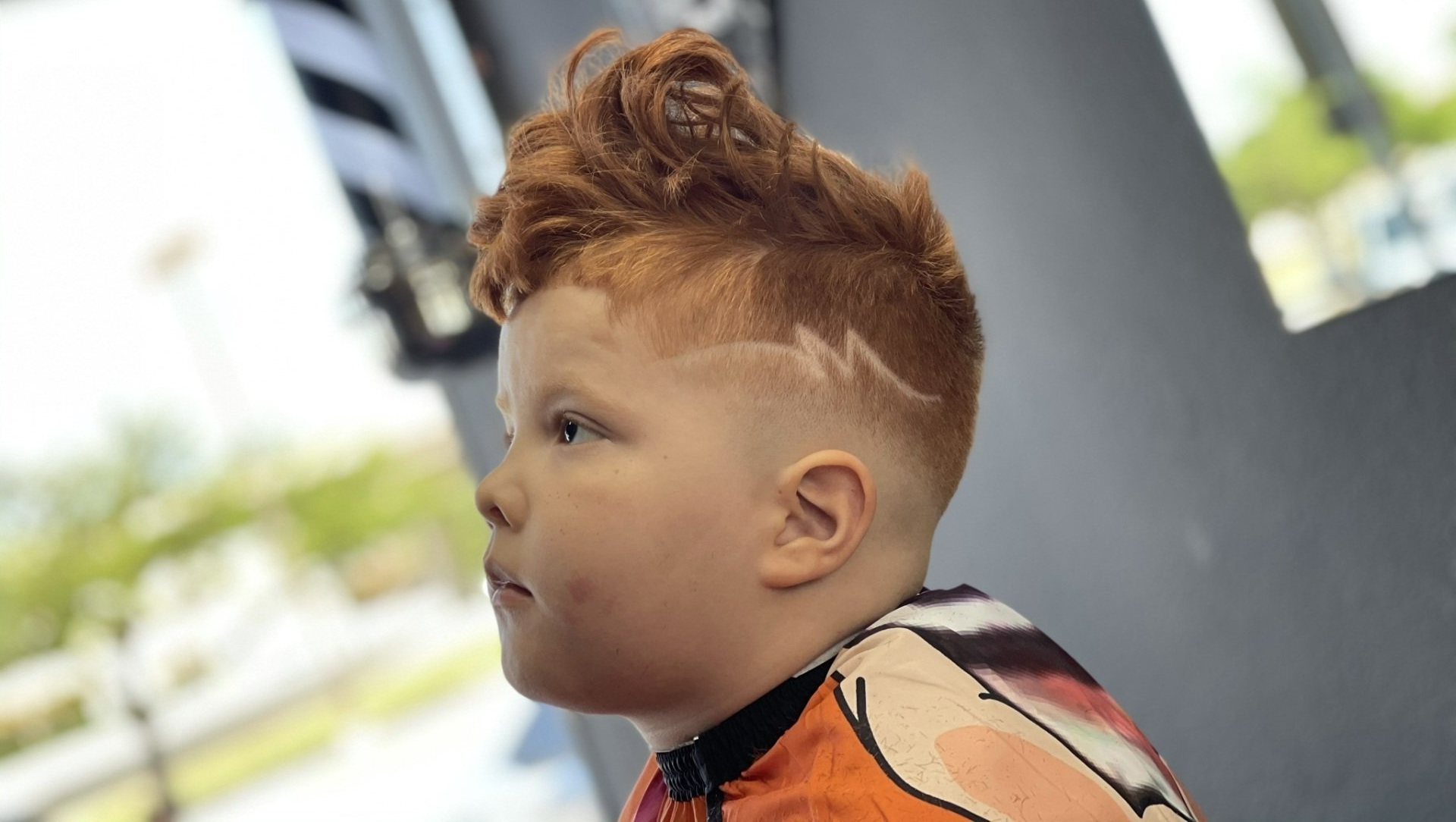 Close-up of a child with curly red hair in an orange shirt, looking left indoors.