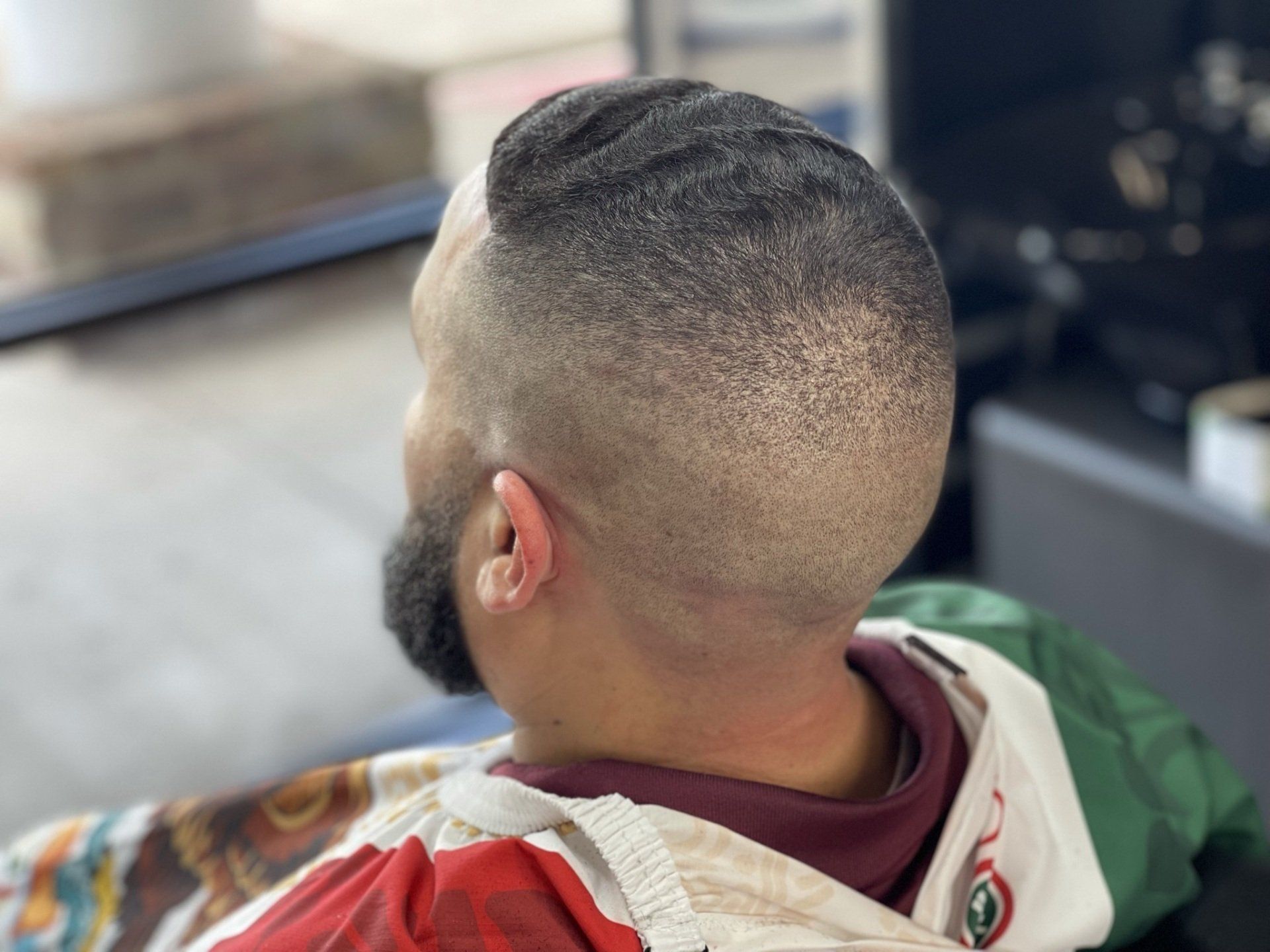Man with a close-cropped fade haircut and beard, viewed from behind in a barber chair, indoors