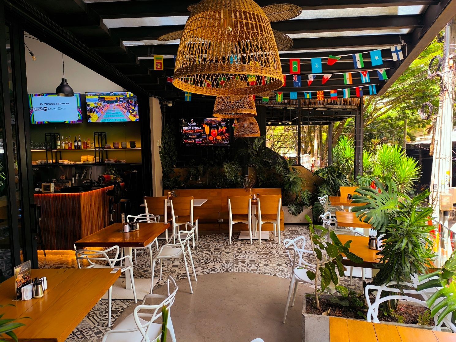 Terraza de restaurante al aire libre con mesas de madera, luces colgantes, televisores y banderas de colores.
