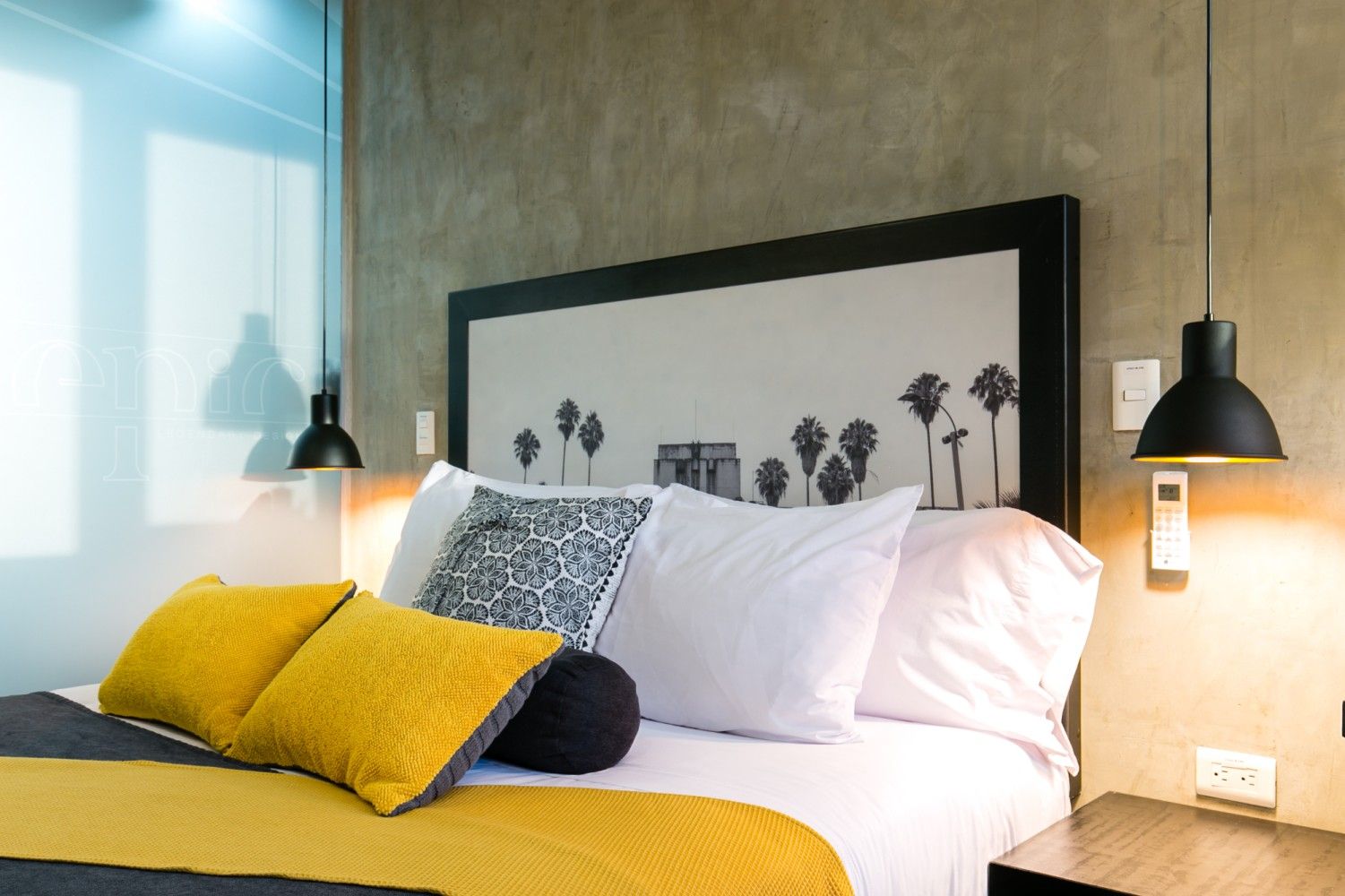 Dormitorio moderno con una colcha amarilla, almohadas blancas, lámparas colgantes negras y un cuadro enmarcado con una palmera.