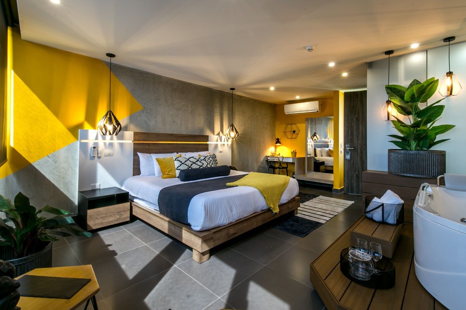 Habitación de hotel moderna con cama, pared decorativa amarilla, zona de estar y plantas.