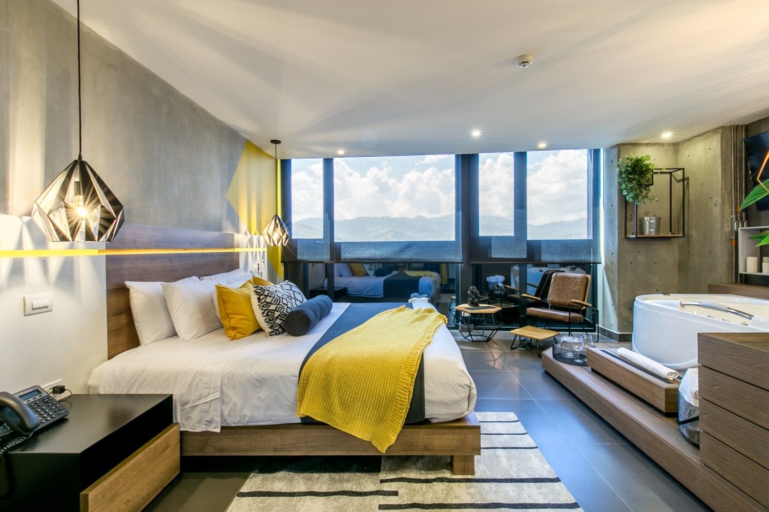 Habitación de hotel moderna con una cama grande, manta amarilla, zona de estar y ventanales del suelo al techo con vistas a la ciudad.