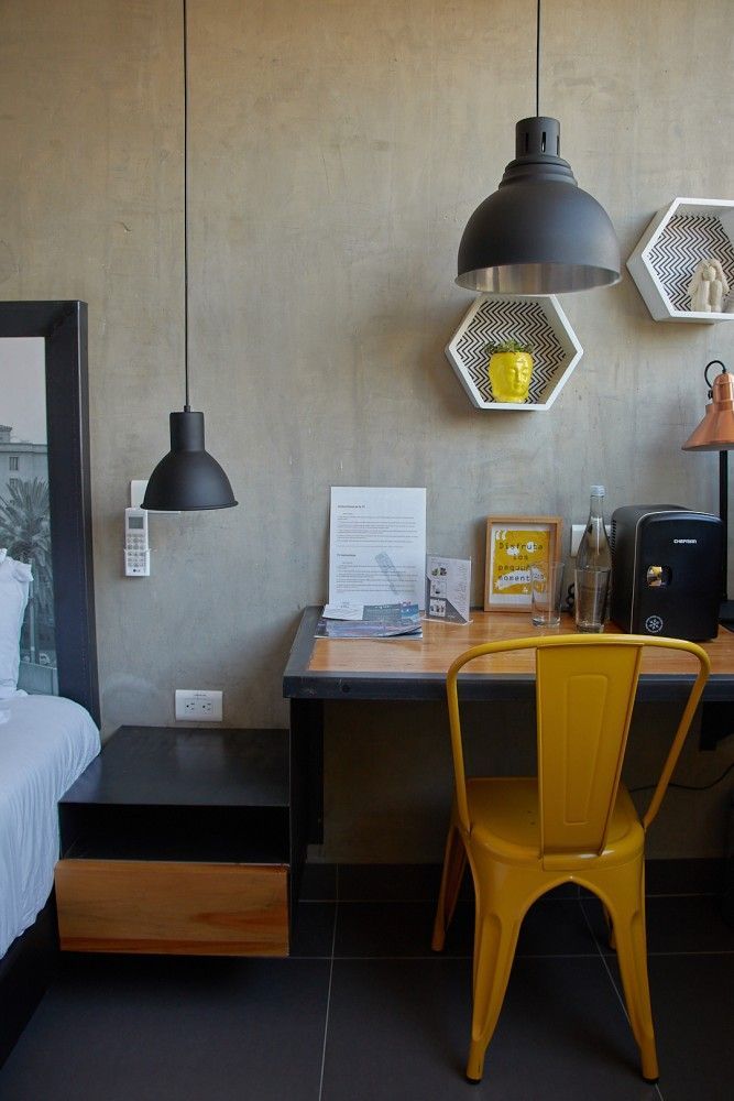 Escritorio pequeño y moderno con silla amarilla, lámpara colgante negra y estante hexagonal en la pared, sobre una pared gris.