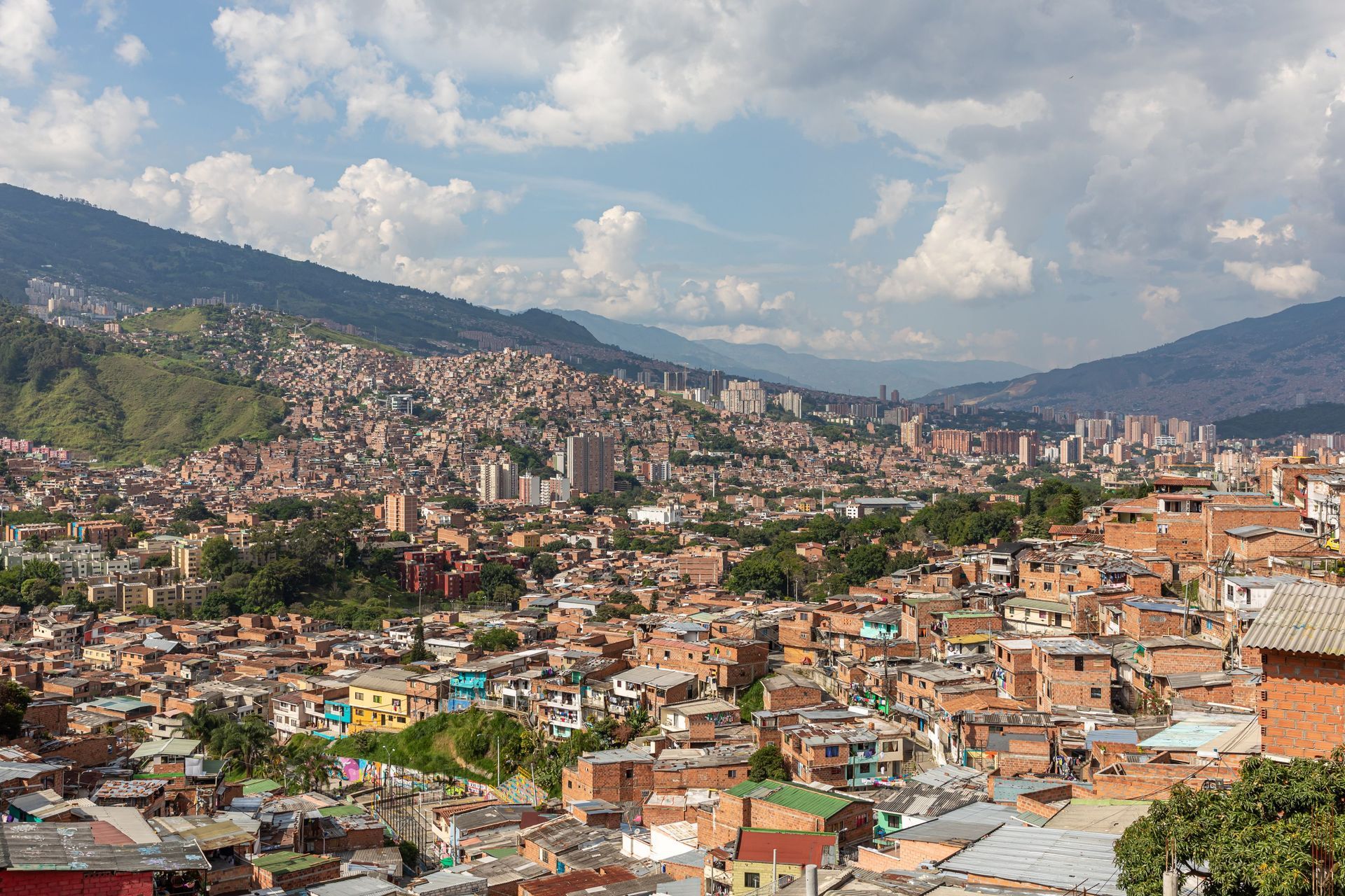 Barrio denso en la ladera de una colina, con casas coloridas bajo montañas verdes y cielo nublado.
