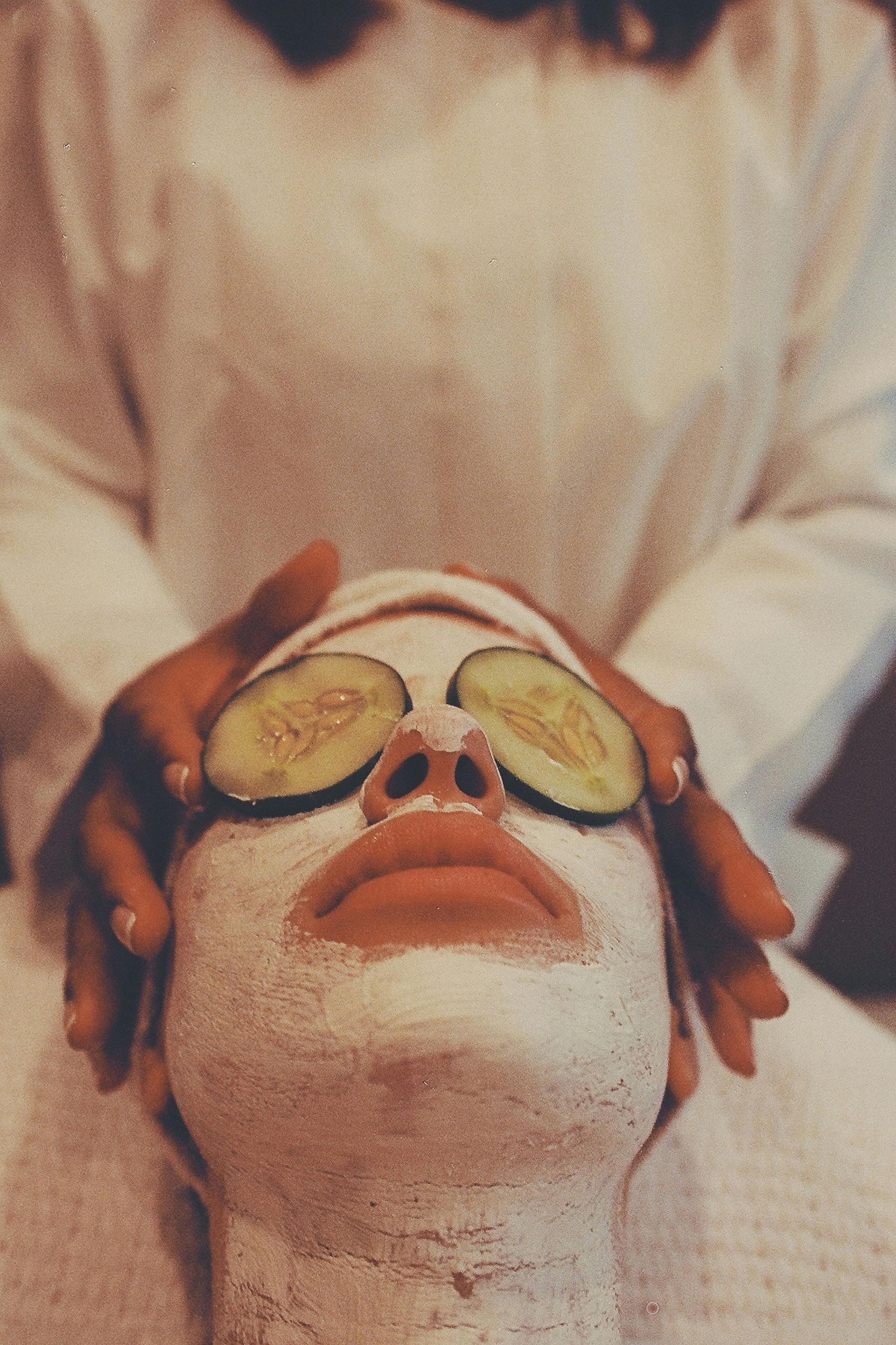 Persona recibiendo una mascarilla facial con rodajas de pepino sobre los ojos y manos que le dan un masaje en la cara.