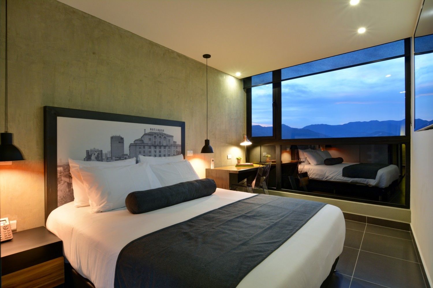 Dormitorio moderno con una cama grande, lámparas de noche y vistas a la ciudad a través de ventanales que van del suelo al techo al atardecer.