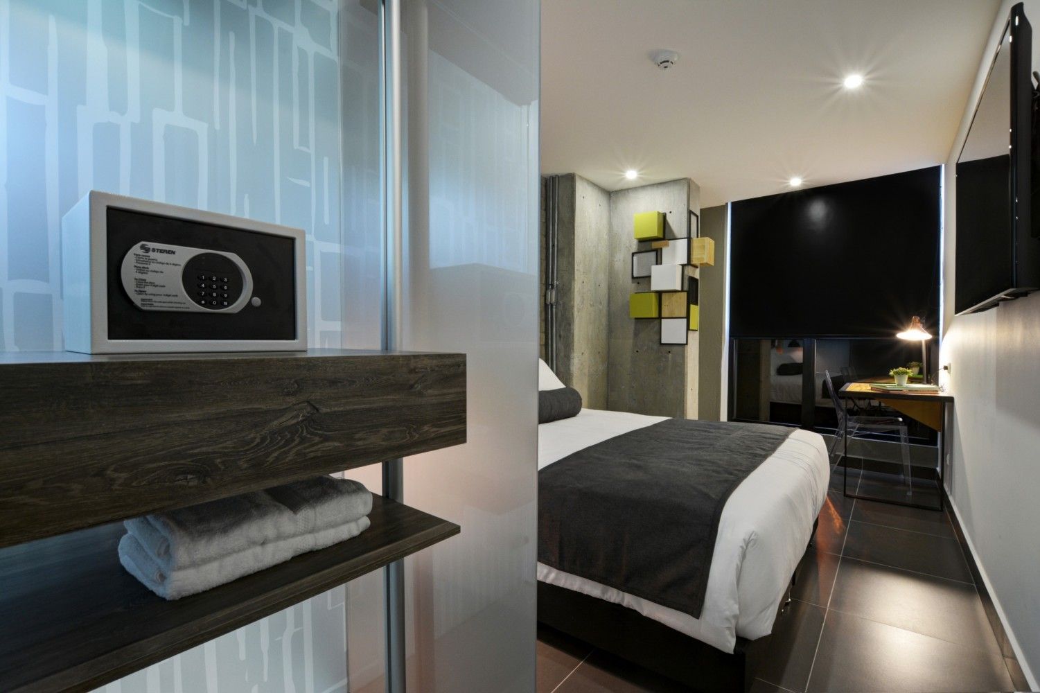 Habitación de hotel moderna con cama, escritorio, caja fuerte y detalles en madera oscura.