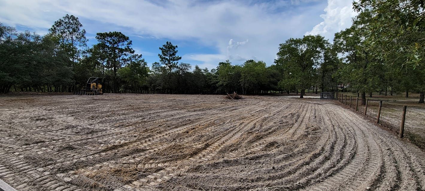 A cleared dirt field with tire tracks, trees in the background, and a fence on the right. Blue sky and clouds above.