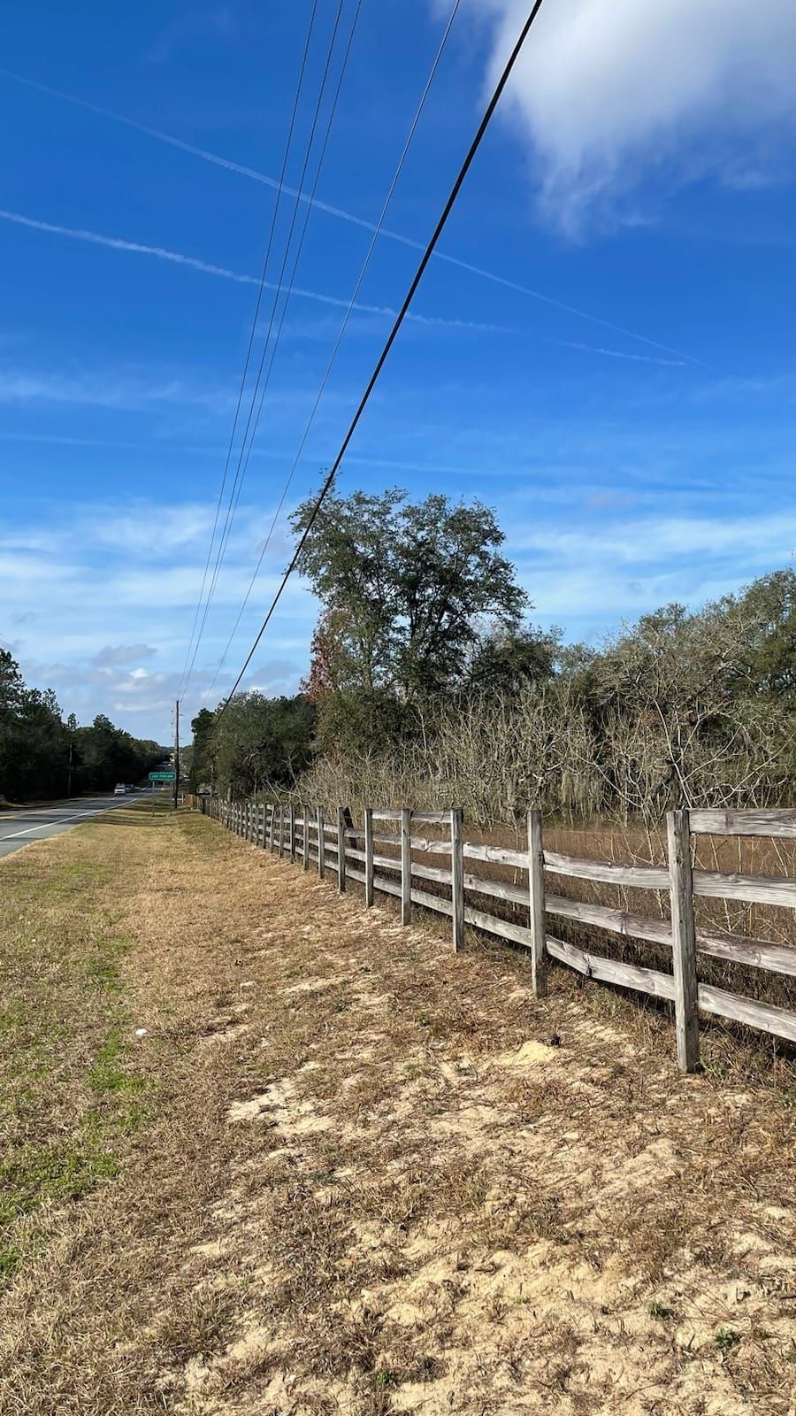 A fence and dried grass in a field, power lines with birds, trees under a blue sky.