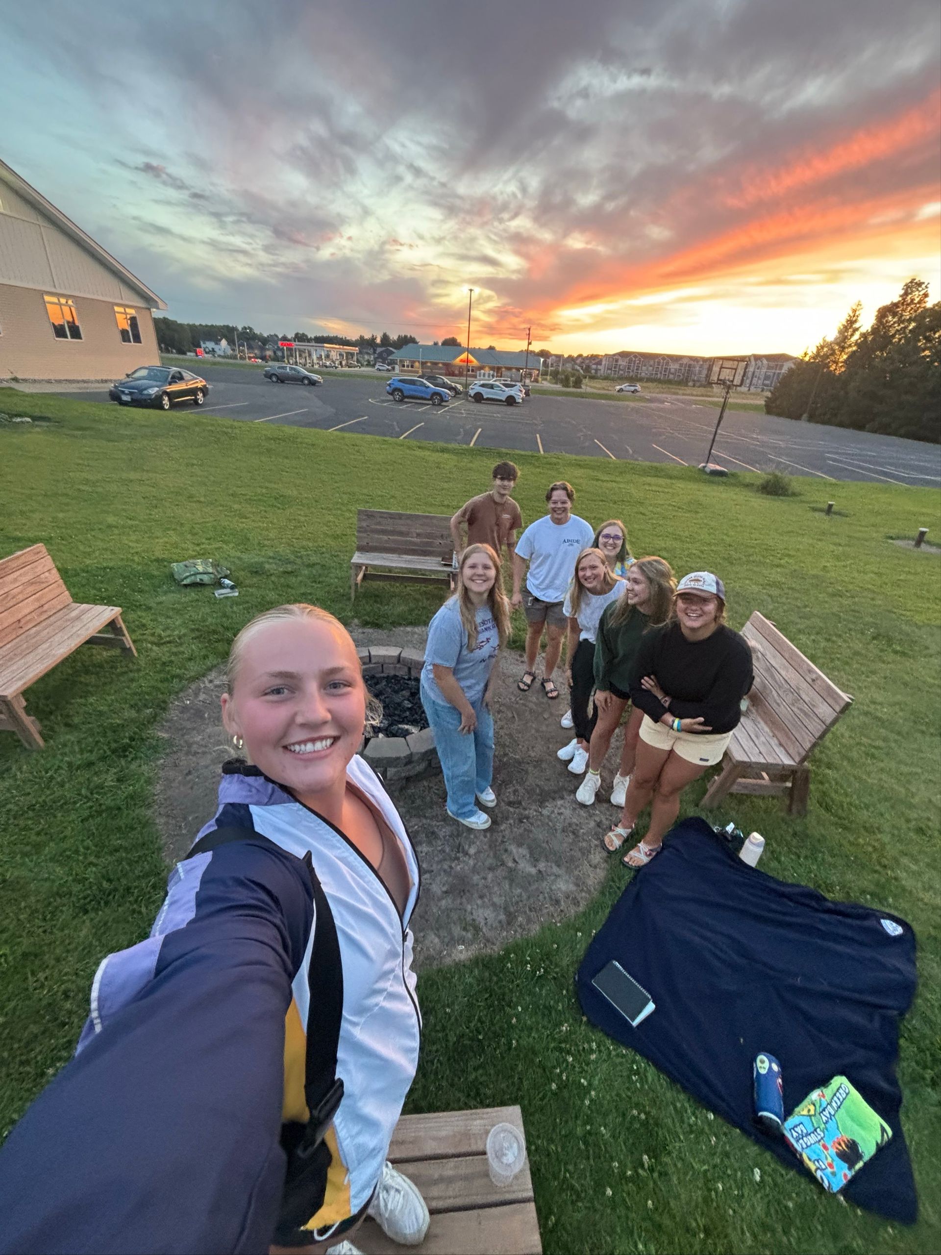 Group of people by a fire pit, taking a selfie during a sunset.