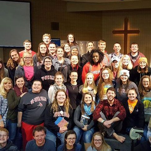 Group of people gathered in a room, some smiling, with a cross in the background.