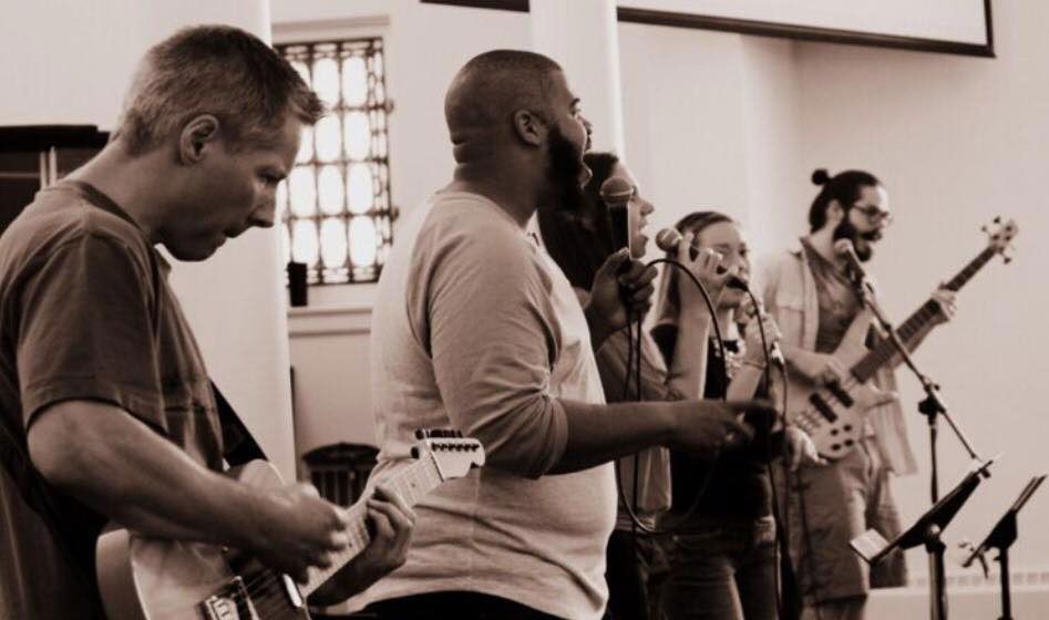 Band performing in a well-lit indoor space, singing into microphones, guitarist, bassist, and background singers present.