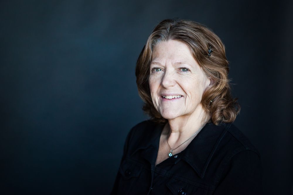 Woman with brown hair, smiling, wearing a black shirt, in front of a dark blue background.