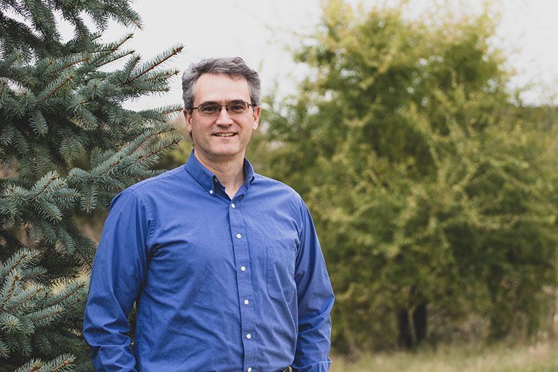 Man in blue shirt and glasses, smiling, standing outdoors near trees.