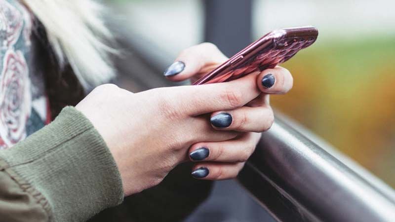 Person holding a pink phone with dark gray nail polish, outdoors.