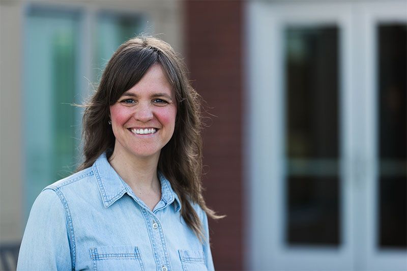 Woman in denim shirt smiles in front of a building with glass doors.