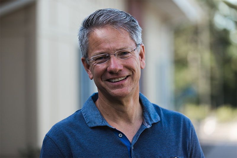 Man with gray hair and glasses smiling outdoors in a blue shirt.