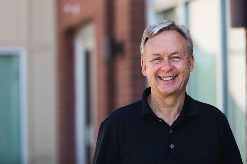 Man with gray hair smiling outside a building, wearing a black shirt.