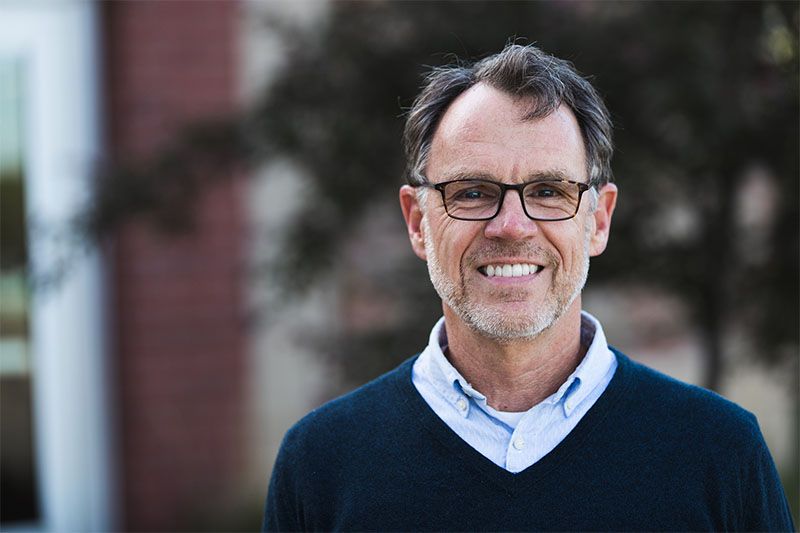Man with glasses smiling, wearing a blue sweater and light blue shirt, outdoors near a brick building.