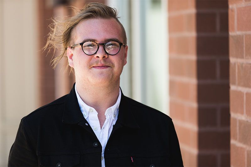Man with glasses and a button-down shirt stands outside near a brick wall, smiling.