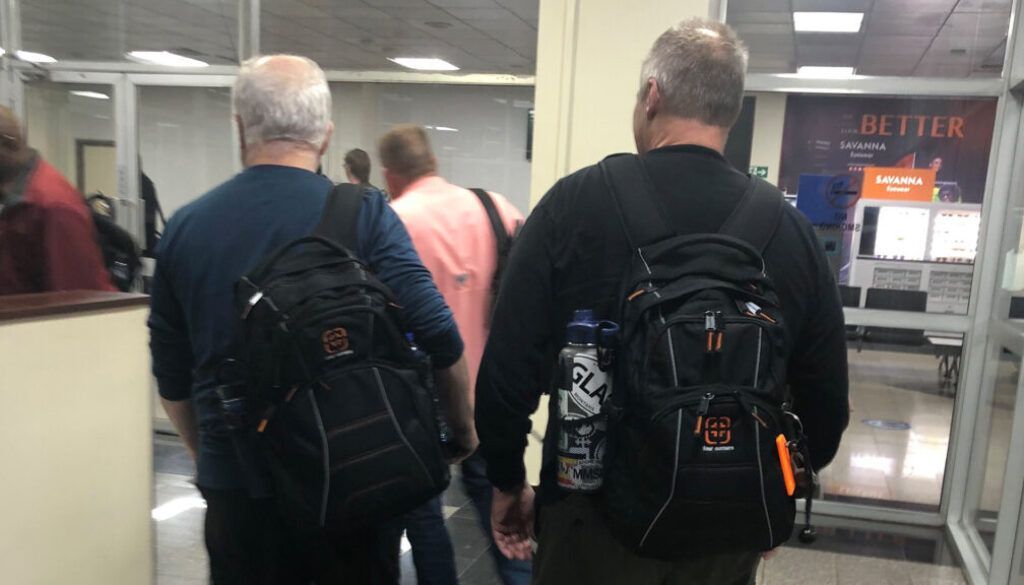Artboard 1 Two men with backpacks walking down a hallway, other people visible in the background.
