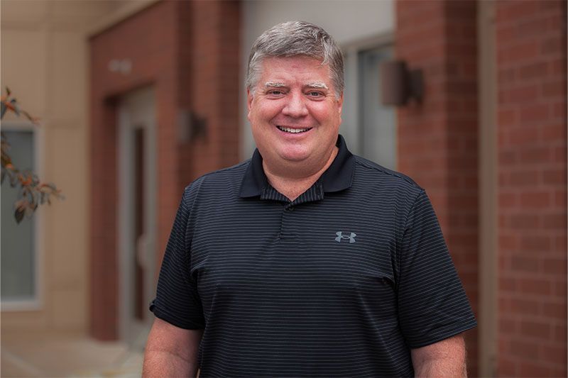 Man in black polo shirt smiles in front of building with brick accents.